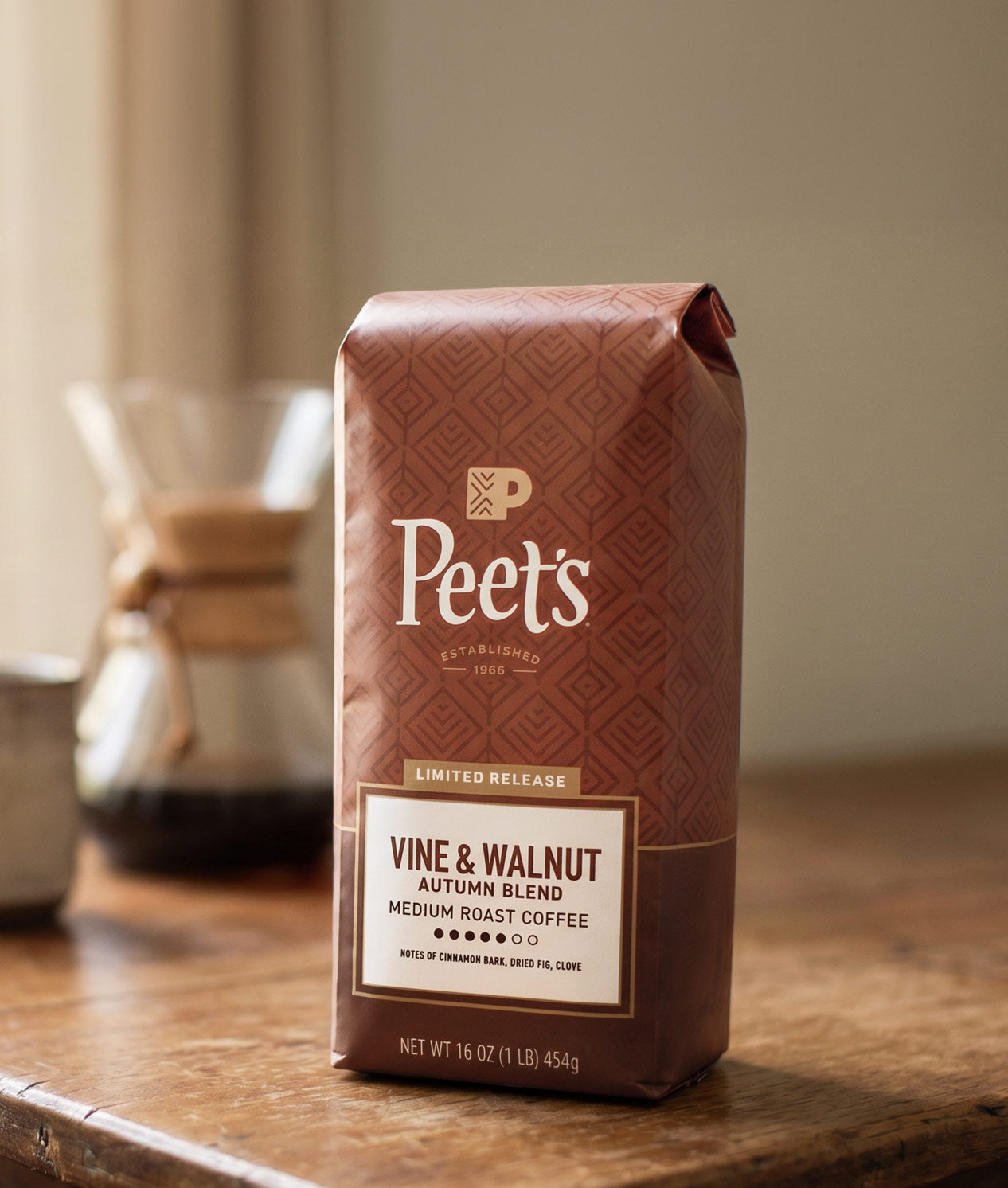 A bag of Peet's Vine & Walnut Autumn Blend medium roast coffee sits on a wooden table with a blurred coffee maker in the background.